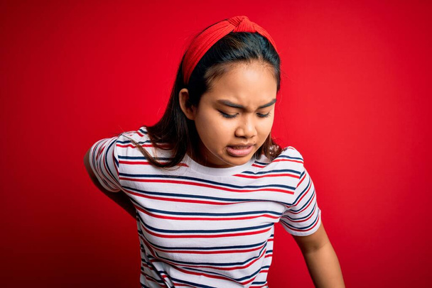Young beautiful asian girl wearing casual striped t-shirt over isolated red background Suffering of backache, touching back with hand, muscular pain - Foto, afbeelding