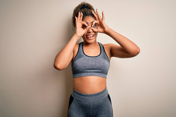 Young african american sportswoman doing sport wearing sportswear over white background doing ok gesture like binoculars sticking tongue out, eyes looking through fingers. Crazy expression. - Фото, зображення
