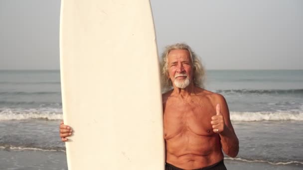 Viejo hombre maduro con tabla de surf en la playa mostrando los pulgares hacia arriba
 - Imágenes, Vídeo
