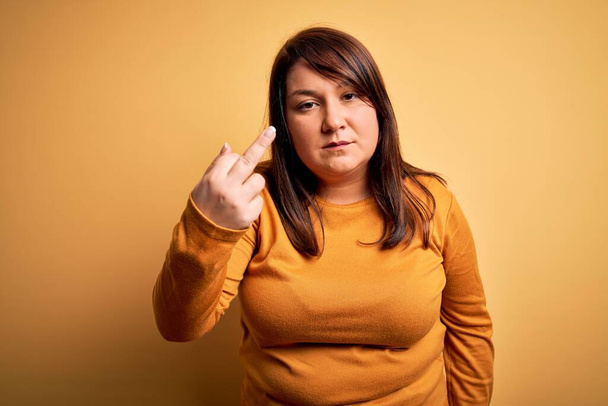 Beautiful brunette plus size woman wearing casual sweater over isolated yellow background Showing middle finger, impolite and rude fuck off expression - Photo, Image