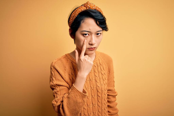 Joven hermosa chica asiática vistiendo suéter casual y diadema de pie sobre fondo amarillo Señalando a la vista observando gesto, expresión sospechosa
 - Foto, imagen