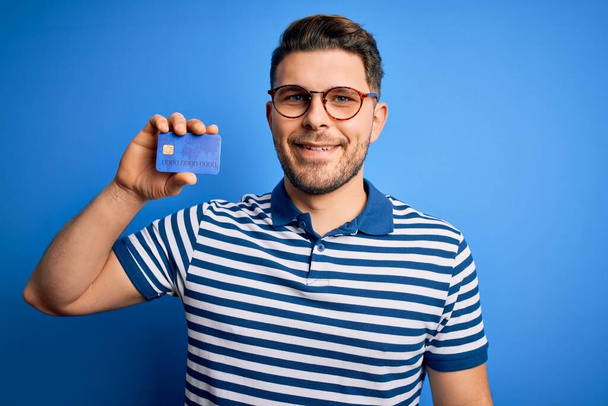 Młody człowiek z niebieskimi oczami, noszący okulary i trzymający kartę kredytową na niebieskim tle ze szczęśliwą twarzą stojącą i uśmiechniętą z pewnym siebie uśmiechem pokazującym zęby - Zdjęcie, obraz