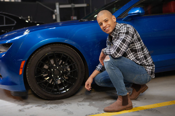 Cheerful attractive African man smiling to the camera while working at his garage with his new sports car - Photo, Image