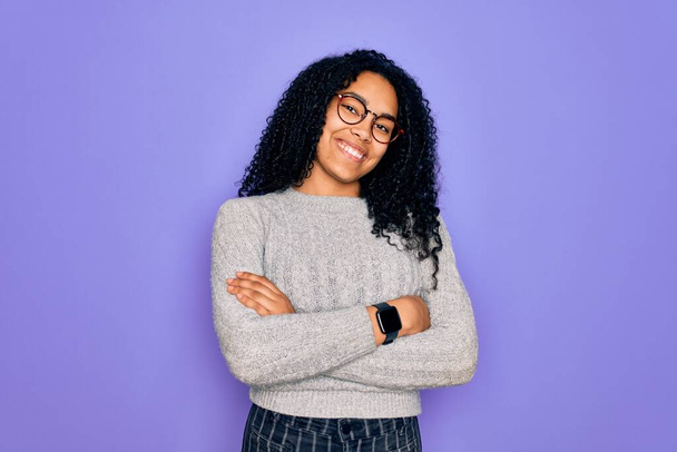 Jeune femme afro-américaine portant un pull décontracté et des lunettes sur fond violet heureux visage souriant avec les bras croisés regardant la caméra. Personne positive
. - Photo, image
