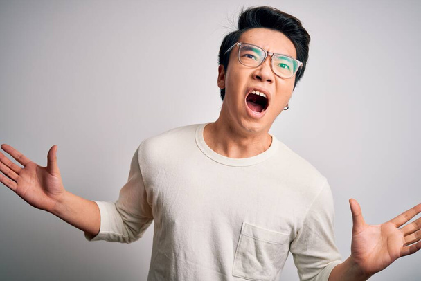 Giovane bell'uomo cinese indossa casual t-shirt e occhiali su sfondo bianco pazzo e pazzo urlando e urlando con espressione aggressiva e braccia alzate. Concetto di frustrazione
. - Foto, immagini