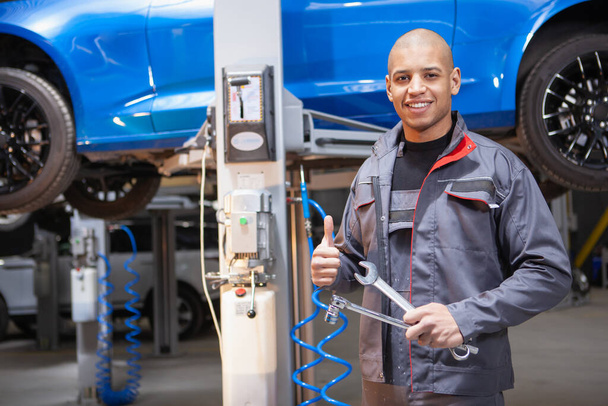 Feliz joven técnico de coches mostrando pulgares hacia arriba mientras se repara un coche en el taller
 - Foto, Imagen