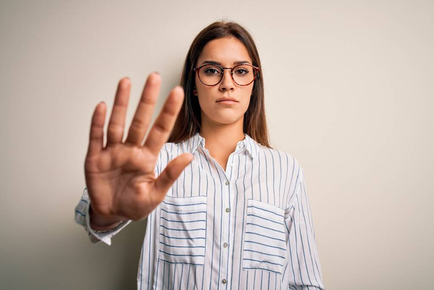 Young beautiful brunette woman wearing casual shirt and glasses over white background doing stop sing with palm of the hand. Warning expression with negative and serious gesture on the face. - Photo, Image