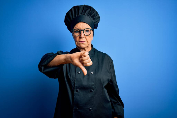 Старший красивый седой шеф-повар женщина в форме плита и шляпа на голубом фоне выглядит несчастным и сердитым показывая отказ и негативный с большим пальцем вниз жест. Плохое выражение
. - Фото, изображение