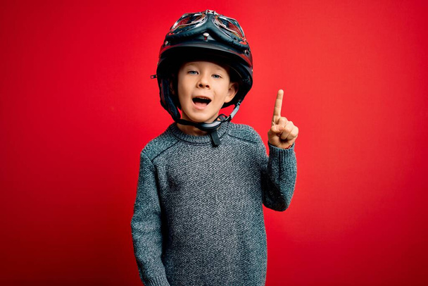 Kleines kaukasisches Kind mit Motorradhelm und Brille über rotem Hintergrund, das mit erhobenem Zeigefinger auf eine erfolgreiche Idee hinweist. Aufgeregt und glücklich. Nummer eins. - Foto, Bild