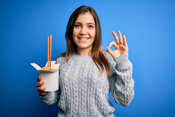 Jeune femme mangeant des nouilles asiatiques de la boîte à emporter à l'aide de baguette sur fond bleu faisant signe ok avec les doigts, excellent symbole
 - Photo, image