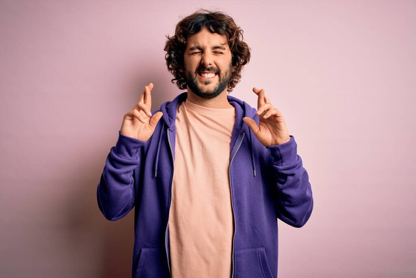 Young handsome sporty man with beard wearing casual sweatshirt over pink background gesturing finger crossed smiling with hope and eyes closed. Luck and superstitious concept. - Zdjęcie, obraz
