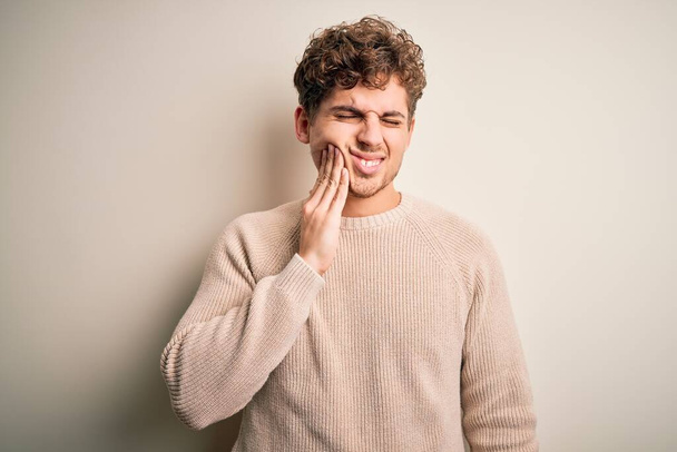 Mladý blonďatý pohledný muž s kudrnatými vlasy na sobě ležérní svetr přes bílé pozadí dotýká úst s rukou s bolestivým výrazem, protože zuby nebo zubní onemocnění na zubech. Zubař - Fotografie, Obrázek