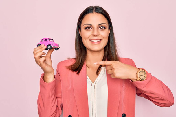 Jovem mulher de negócios bonita morena segurando pequeno carro brinquedo sobre fundo rosa Sorrindo feliz apontando com a mão e o dedo
 - Foto, Imagem