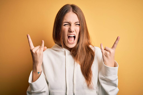 Young beautiful redhead sporty woman wearing sweatshirt over isolated yellow background shouting with crazy expression doing rock symbol with hands up. Music star. Heavy concept. - Fotó, kép