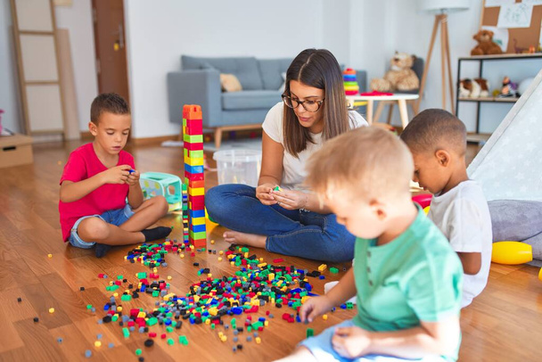 Joven hermosa maestra y niños pequeños jugando con bloques de construcción alrededor de un montón de juguetes en el jardín de infantes
 - Foto, Imagen