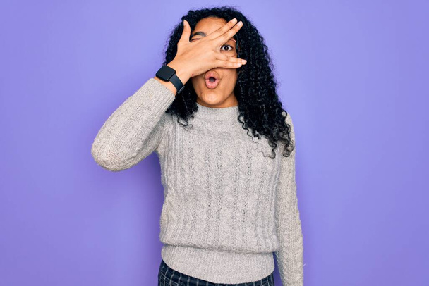 Mujer afroamericana joven que usa suéter casual y gafas sobre fondo púrpura asomándose en la cara y los ojos de cobertura de choque con la mano, mirando a través de los dedos con expresión embarazosa
. - Foto, Imagen