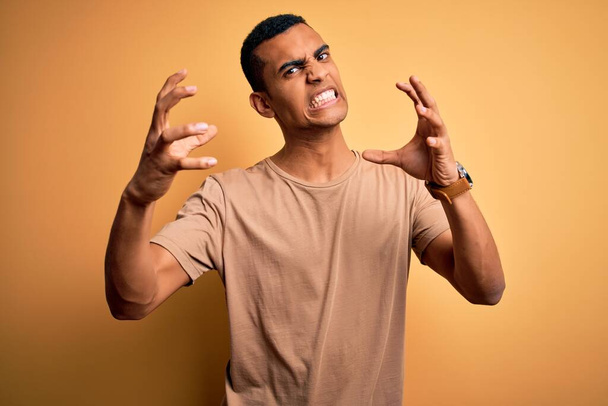Mladý pohledný africký Američan v neformálním tričku, stojící nad žlutým pozadím, křičící frustrovaný vztekem, ruce snažící se uškrtit, ječící vzteky - Fotografie, Obrázek