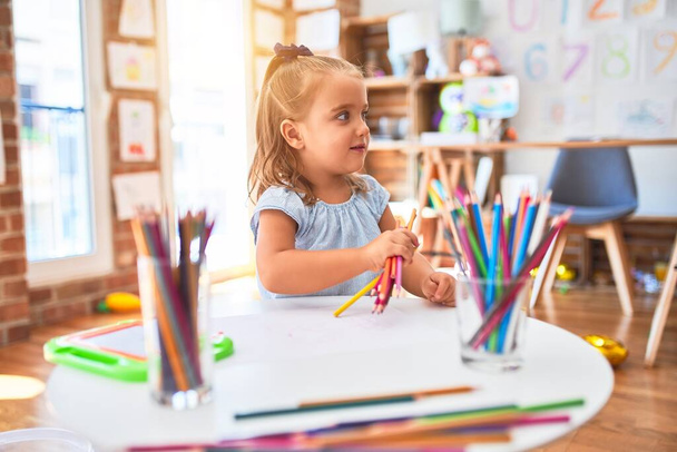 Молодая красивая блондинка девочка наслаждается игрой школы с игрушками в детском саду, улыбаясь счастливая картина с карандашом цвета дома
 - Фото, изображение