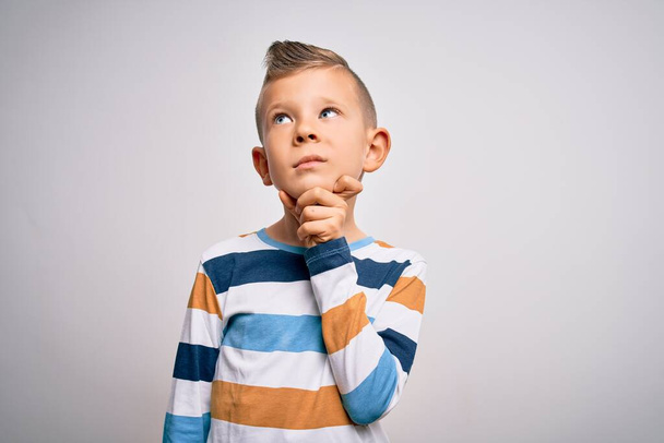 Young little caucasian kid with blue eyes standing wearing striped shirt over isolated background with hand on chin thinking about question, pensive expression. Smiling with thoughtful face. Doubt concept. - Photo, Image