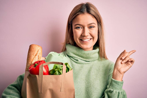Young beautiful blonde girl holding fresh groceries paper bag over pink isolated background very happy pointing with hand and finger to the side - Photo, Image