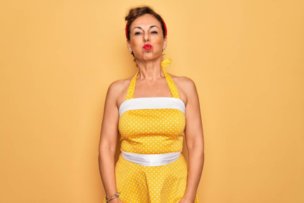 Seniorinnen mittleren Alters tragen ein Retro-Kleid im 50er-Jahre-Stil über gelbem Hintergrund mit aufgeblasenen Wangen und lustigem Gesicht. Mund mit Luft aufgeblasen, verrückter Ausdruck. - Foto, Bild