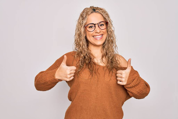 Young beautiful blonde woman wearing casual sweater and glasses over white background success sign doing positive gesture with hand, thumbs up smiling and happy. Cheerful expression and winner gesture. - Photo, Image