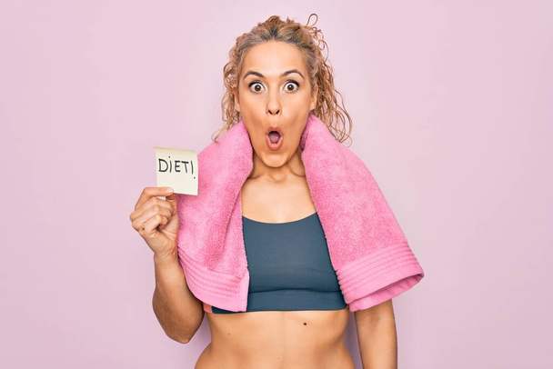 Beautiful blonde sporty woman doing sport wearing towel holding reminder with diet message scared and amazed with open mouth for surprise, disbelief face - Photo, Image