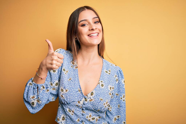 Young beautiful brunette woman wearing casual floral t-shirt standing over yellow background doing happy thumbs up gesture with hand. Approving expression looking at the camera showing success. - Фото, зображення
