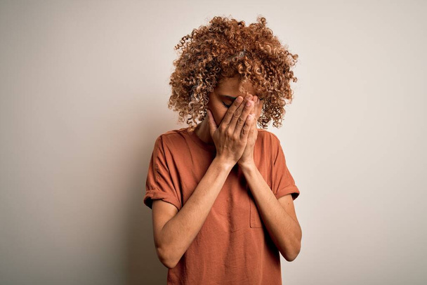 Όμορφη Αφροαμερικανή γυναίκα με σγουρά μαλλιά που φοράει casual t-shirt πάνω από λευκό φόντο με λυπητερή έκφραση που καλύπτει το πρόσωπο με τα χέρια ενώ κλαίει. Έννοια κατάθλιψης. - Φωτογραφία, εικόνα