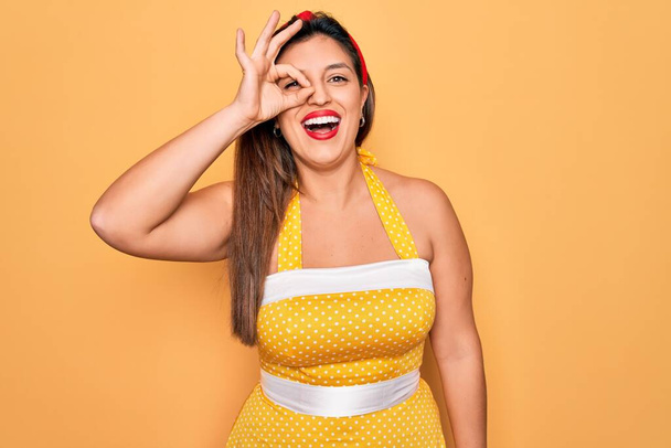 Joven hispano pin up mujer usando moda sexy estilo de los años 50 sobre fondo amarillo haciendo buen gesto con la mano sonriente, ojo mirando a través de los dedos con cara feliz
. - Foto, Imagen