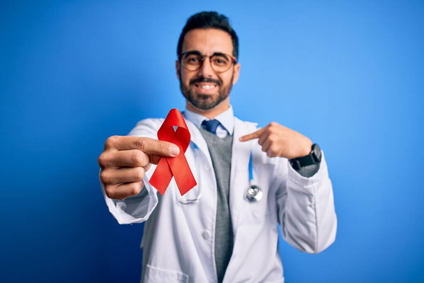Young handsome doctor man with beard wearing stethoscope holding red hiv ribbon with surprise face pointing finger to himself - Foto, immagini