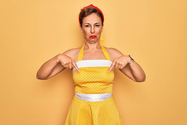 Orta yaşlı bir kadın, 50 'li yaşlarda, sarı arka planda eski tarz bir elbise giymiş üzgün ve üzgün görünüyor, parmaklarıyla yönü gösteriyor, mutsuz ve depresif.. - Fotoğraf, Görsel