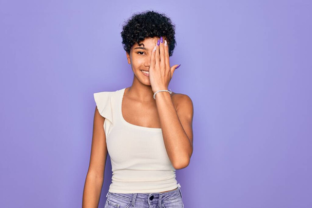 Młoda, piękna afrykańska afro kobieta ubrana w luźny t-shirt na fioletowym tle pokrywającym jedno oko ręką, pewny siebie uśmiech na twarzy i niespodziewane emocje. - Zdjęcie, obraz