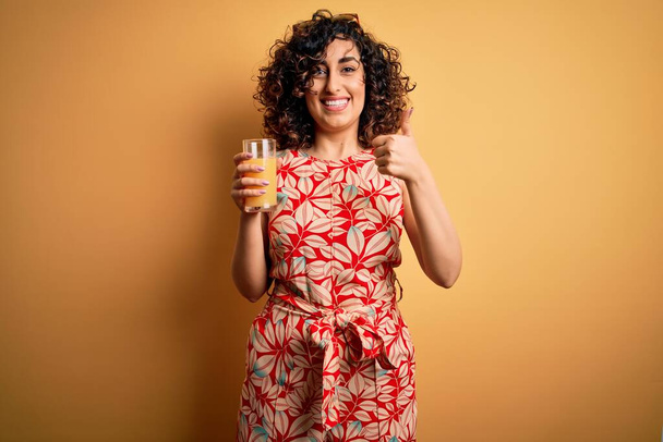 Young beautiful curly arab woman drinking glass of healthy orange juice over yellow background happy with big smile doing ok sign, thumb up with fingers, excellent sign - Fotó, kép