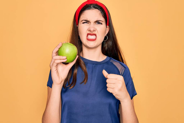 Młoda piękna dziewczyna jedząc świeże organiczne zdrowe zielone jabłko na żółtym tle zirytowany i sfrustrowany krzycząc ze złości, szalony i krzycząc z podniesionymi rękami, koncepcja złości - Zdjęcie, obraz