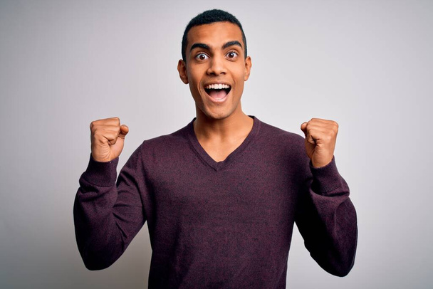 Young handsome african american man wearing casual sweater over white background celebrating surprised and amazed for success with arms raised and open eyes. Winner concept. - Foto, imagen