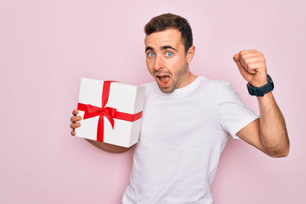 Young handsome man with blue eyes holding birthday present over isolated pink background screaming proud and celebrating victory and success very excited, cheering emotion - Foto, imagen