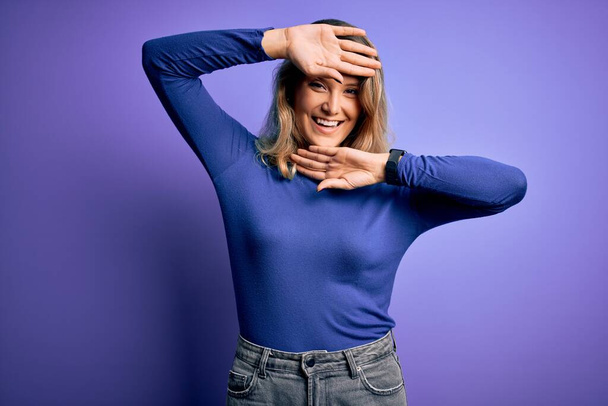 Nuori kaunis blondi nainen yllään rento t-paita yli eristetty violetti tausta Hymyilevä iloinen pelaa kurkistaa boo kädet osoittavat kasvot. Yllätys ja poistuminen
 - Valokuva, kuva