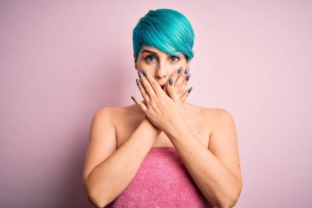 Mladá žena s modrými módními vlasy nosí ručník po sprše přes růžové pozadí šokován pokrývající ústa s rukama pro omyl. Tajný koncept. - Fotografie, Obrázek