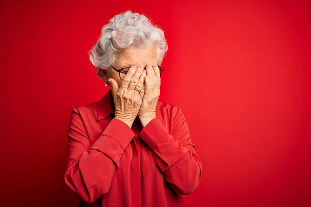 Starší krásná šedovlasá žena v neformální košili a brýlích na červeném pozadí se smutným výrazem zakrývajícím tvář rukama při pláči. Koncept deprese. - Fotografie, Obrázek