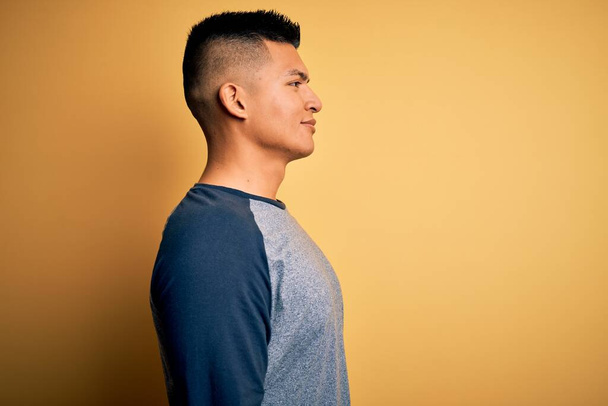 Young handsome latin man wearing casual t-shirt standing over yellow background looking to side, relax profile pose with natural face with confident smile. - Photo, Image