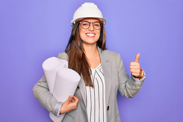 Молодая латиноамериканская архитектор женщина в шляпе строителя и держа чертежи на желтом фоне счастливы с большой улыбкой делает хорошо знак, палец вверх пальцами, отличный знак
 - Фото, изображение
