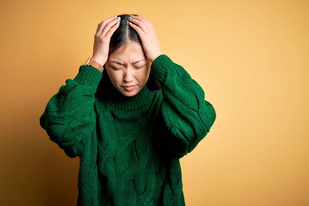 Jovem bela mulher asiática vestindo camisola de inverno verde sobre fundo isolado amarelo sofrendo de dor de cabeça desesperada e estressada porque dor e enxaqueca. Mãos na cabeça
. - Foto, Imagem