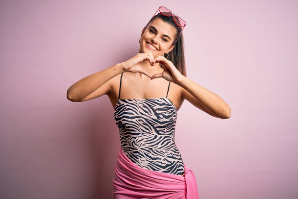 Jonge mooie brunette vrouw op vakantie dragen badpak over roze achtergrond glimlachen in liefde doen hart symbool vorm met de handen. Romantisch concept. - Foto, afbeelding