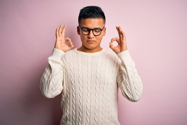 Joven hombre latino guapo usando suéter casual blanco y gafas sobre fondo rosa relajarse y sonreír con los ojos cerrados haciendo gesto de meditación con los dedos. Concepto de yoga
. - Foto, Imagen
