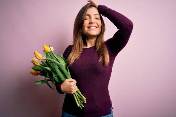 Mulher loira jovem segurando buquê romântico de flores de tulipas amarelas sobre fundo rosa sorrindo confiante tocando o cabelo com mão para cima gesto, posando atraente e elegante
 - Foto, Imagem