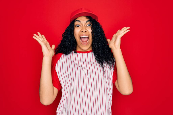 Young african american curly sportswoman wearing baseball cap and striped t-shirt celebrating crazy and amazed for success with arms raised and open eyes screaming excited. Winner concept - Photo, Image