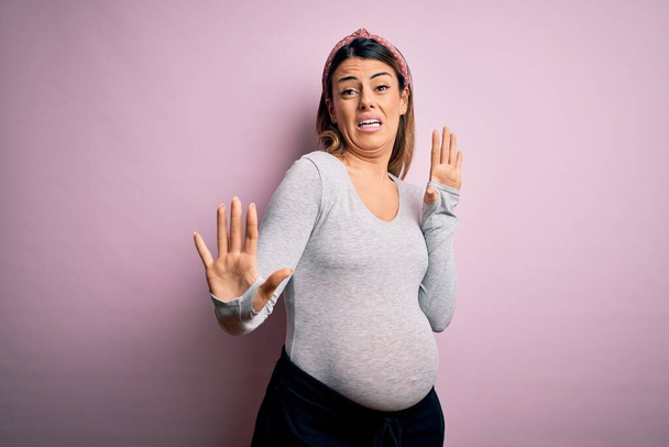 Νεαρή όμορφη μελαχρινή γυναίκα έγκυος περιμένει μωρό πάνω από απομονωμένο ροζ φόντο φοβισμένος και τρομοκρατημένος από την έκφραση του φόβου σταματήσει χειρονομία με τα χέρια, φωνάζοντας σε κατάσταση σοκ. Έννοια πανικού. - Φωτογραφία, εικόνα