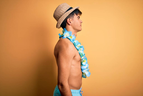 Jeune homme touriste beau en vacances portant des maillots de bain et des fleurs hawaïennes de lei regardant vers le côté, pose de profil de détente avec visage naturel avec sourire confiant
. - Photo, image