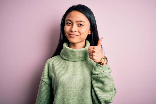Młoda piękna Chinka ubrana w sweter z golfem na odizolowanym różowym tle, robiąca gesty rękami w górę. Zatwierdzanie ekspresji patrząc w aparat pokazujący sukces. - Zdjęcie, obraz